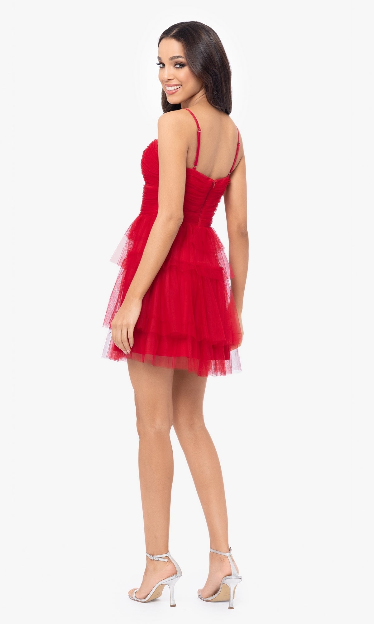 Smiling over her shoulder, a woman wears the Cut-Out Short Ruffled Homecoming Dress 5098BN with spaghetti straps and tiered tulle ruffles, paired with white high-heeled sandals against a plain background.